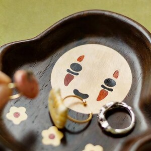 May include: A dark brown wooden tray with a cartoon face design in the center. The tray holds several rings and small flower-shaped decorations. The cartoon face has black eyes, a black mouth, and red cheek markings.