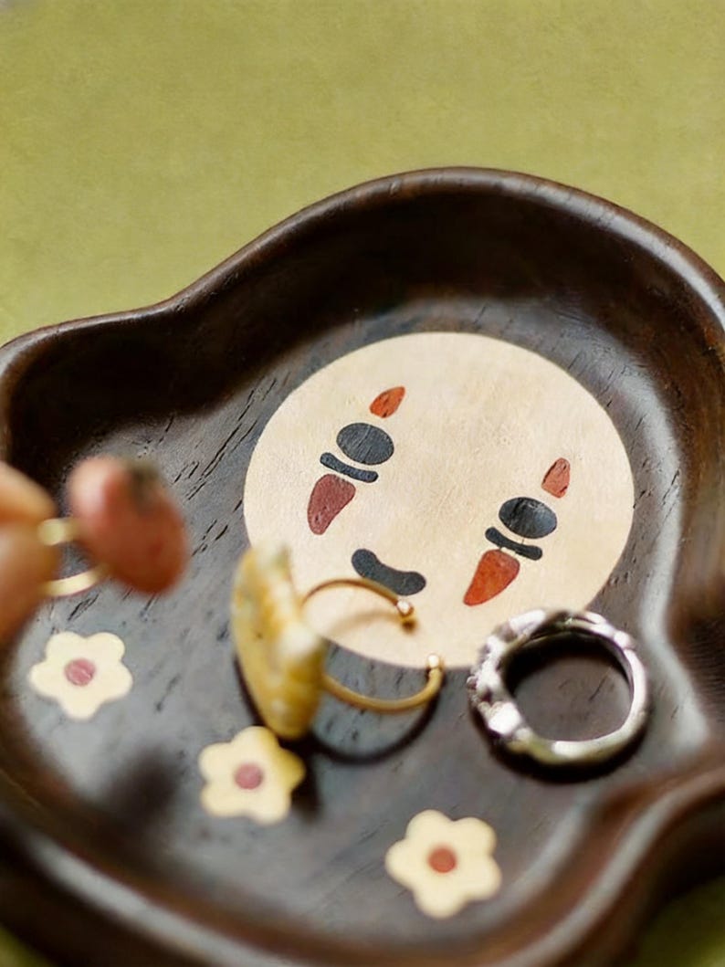 May include: A dark brown wooden tray with a cartoon face design in the center. The tray holds several rings and small flower-shaped decorations. The cartoon face has black eyes, a black mouth, and red cheek markings.