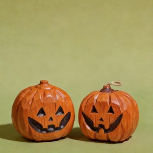 May include: Two hand-carved wooden jack-o'-lantern pumpkins with a natural orange finish. Each pumpkin features a carved face with triangular eyes, a small triangular nose, and a wide, jagged mouth. One pumpkin has a small piece of twine attached.