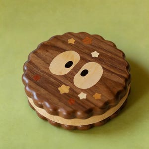 May include: A round wooden box with a whimsical design. The box is made of dark brown wood with a wavy edge and features a cartoon face with light wood eyes and star accents. The box is approximately 4 inches in diameter.