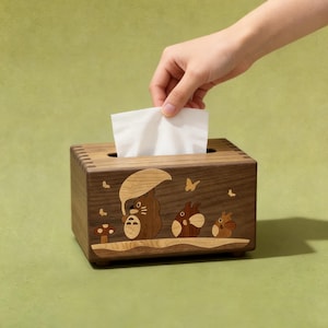 May include: A rectangular wooden tissue box featuring an inlaid design of three cartoon creatures, one holding an umbrella, with a mushroom and butterflies. The box has visible finger joints at the corners and a single white tissue protruding from the top.