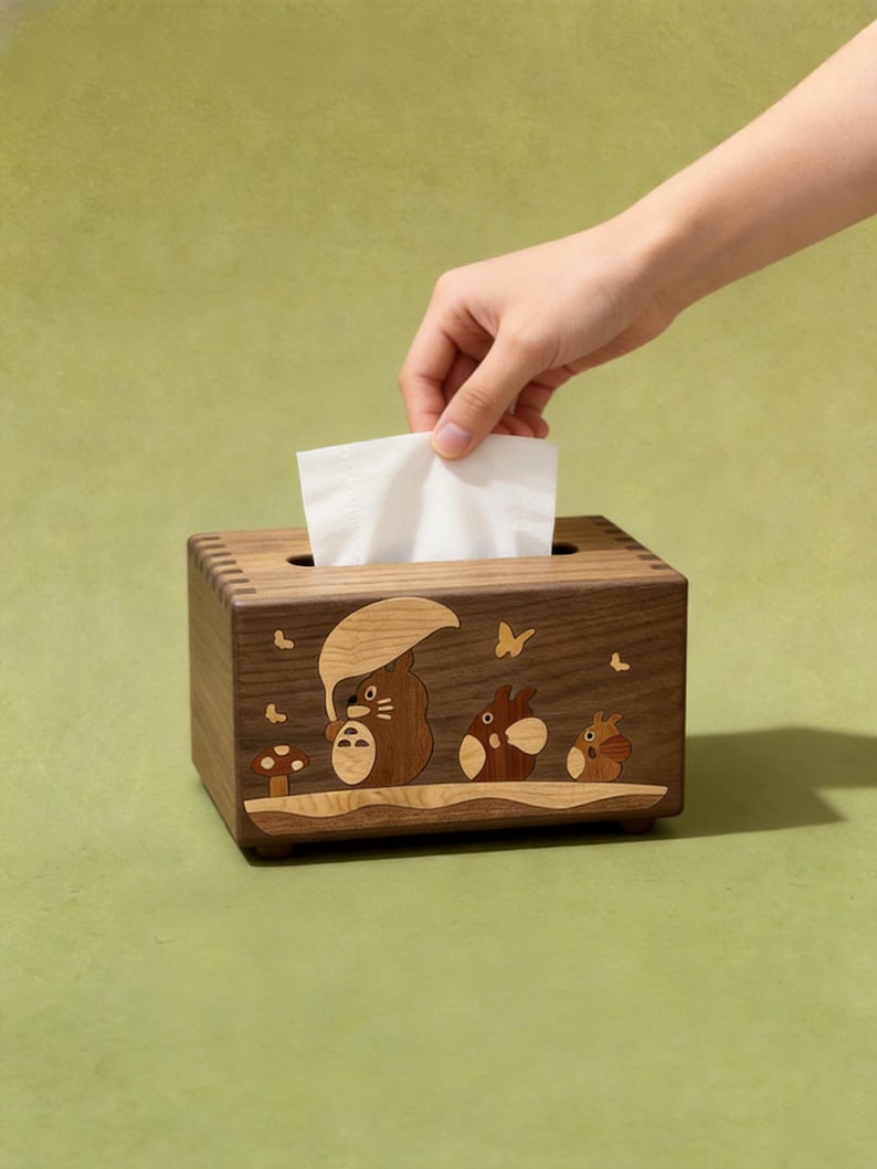 May include: A rectangular wooden tissue box featuring an inlaid design of three cartoon creatures, one holding an umbrella, with a mushroom and butterflies. The box has visible finger joints at the corners and a single white tissue protruding from the top.