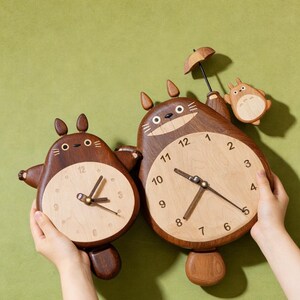 May include: Two wooden wall clocks shaped like cartoon creatures with dark brown bodies and light wood faces. The larger clock features a small figure holding an umbrella, while both have dark brown hands and numerals printed on the light wood face.