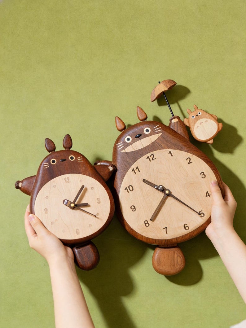 May include: Two wooden wall clocks shaped like cartoon creatures with dark brown bodies and light wood faces. The larger clock features a small figure holding an umbrella, while both have dark brown hands and numerals printed on the light wood face.