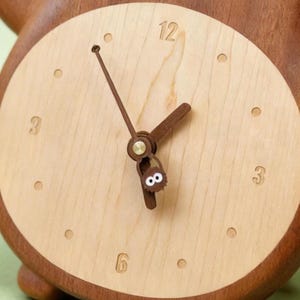 May include: Wooden clock with a light wood face and a dark wood frame. The clock face has the numbers 12, 3, 6, and 9, with small dots marking the other hours. The clock hands are brown, and the second hand has a small, whimsical design.