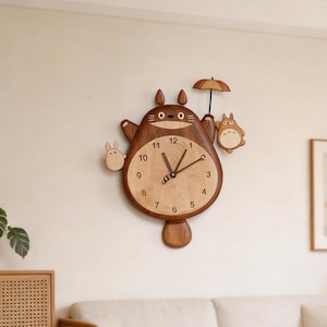 May include: A wooden wall clock shaped like a large brown creature with pointed ears, featuring a light-coloured circular face with black numbers 1 through 12, two dark hands, a small attached umbrella, and two smaller light-coloured figures on the sides.