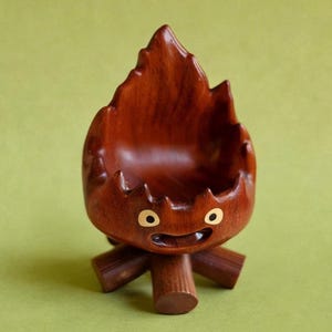 May include: A whimsical, hand-carved wooden bowl in the shape of a stylized flame. The bowl is a rich, dark brown color with a smooth finish. It features a smiling face with two golden eyes and a small mouth. The bowl is supported by three wooden legs.