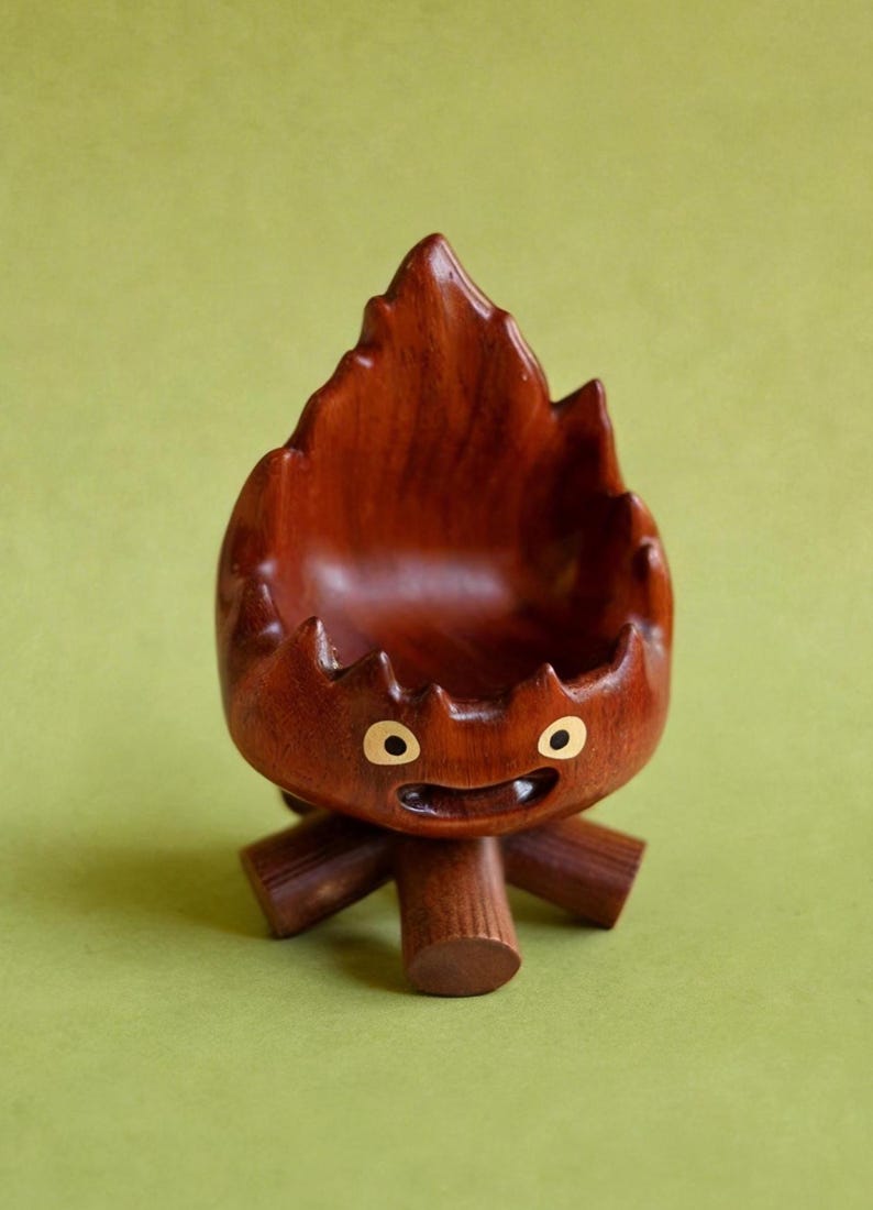 May include: A whimsical, hand-carved wooden bowl in the shape of a stylized flame. The bowl is a rich, dark brown color with a smooth finish. It features a smiling face with two golden eyes and a small mouth. The bowl is supported by three wooden legs.