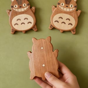 May include: Three wooden cat-shaped magnets. Two magnets are face-up, displaying a smiling cat design with light brown and dark brown wood. The third magnet is held by a hand, showing the back with two small white magnets. The background is a solid green.