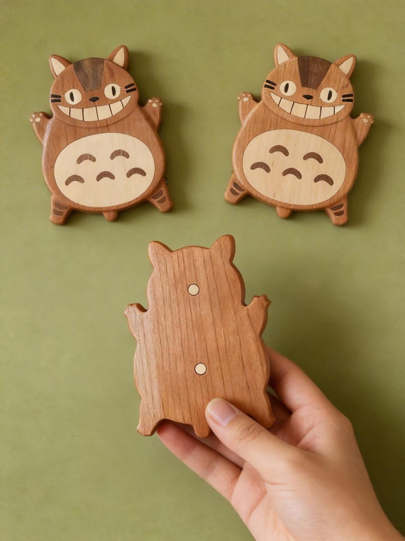 May include: Three wooden cat-shaped magnets. Two magnets are face-up, displaying a smiling cat design with light brown and dark brown wood. The third magnet is held by a hand, showing the back with two small white magnets. The background is a solid green.
