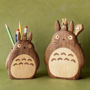 Puede incluir: Dos organizadores de escritorio de madera con forma del personaje Totoro. Los organizadores son marrones con detalles de madera más clara en el vientre. Uno contiene lápices y bolígrafos. El otro tiene un accesorio en forma de cuchara.