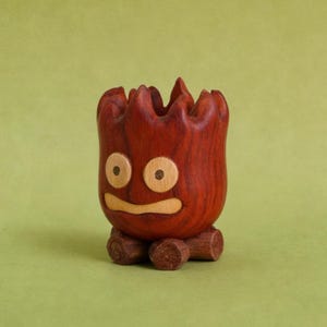 May include: A handcrafted wooden planter with a whimsical face carved into the front. The planter is a rich reddish-brown color with light wood accents for the eyes and mouth. It sits on a base of three small wooden logs.