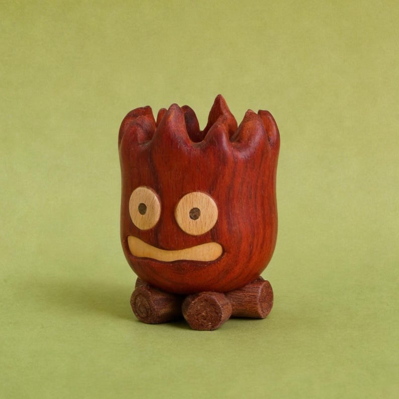 May include: A handcrafted wooden planter with a whimsical face carved into the front. The planter is a rich reddish-brown color with light wood accents for the eyes and mouth. It sits on a base of three small wooden logs.