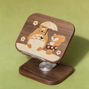May include: A dark wood phone stand featuring an inlaid light wood design of a large cartoon creature holding an umbrella next to a small squirrel sitting on a mushroom. The stand has a silver metal swivel base and a rectangular support panel.