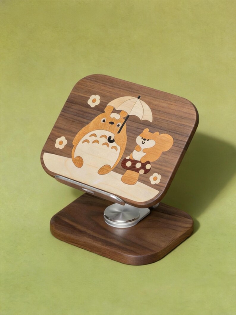 May include: A dark wood phone stand featuring an inlaid light wood design of a large cartoon creature holding an umbrella next to a small squirrel sitting on a mushroom. The stand has a silver metal swivel base and a rectangular support panel.