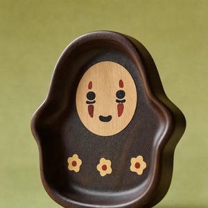 May include: A dark brown wooden tray with a carved design. The tray features a light wood face with black eyes and a red mouth. Three small yellow flowers are carved at the bottom of the tray.