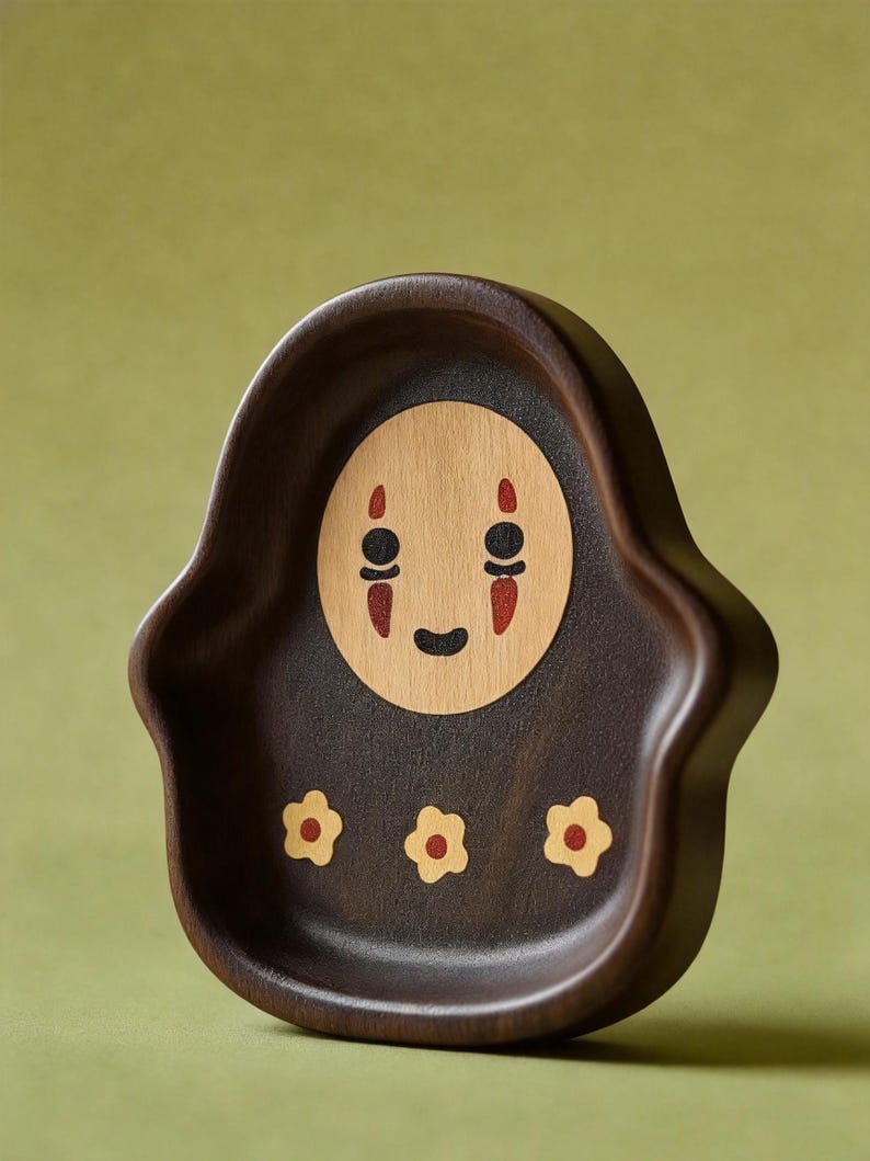 May include: A dark brown wooden tray with a carved design. The tray features a light wood face with black eyes and a red mouth. Three small yellow flowers are carved at the bottom of the tray.