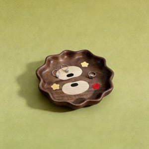 May include: A wooden tray with a bear face design. The tray is brown with light wood and colourful accents. Three rings are placed on the tray. The tray has a wavy edge and is on a green background.