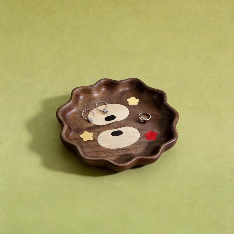 May include: A wooden tray with a bear face design. The tray is brown with light wood and colourful accents. Three rings are placed on the tray. The tray has a wavy edge and is on a green background.