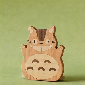 May include: Wooden Totoro figure with a smiling face, paws, and belly markings. The figure is made of light and dark wood tones, with a light green background.