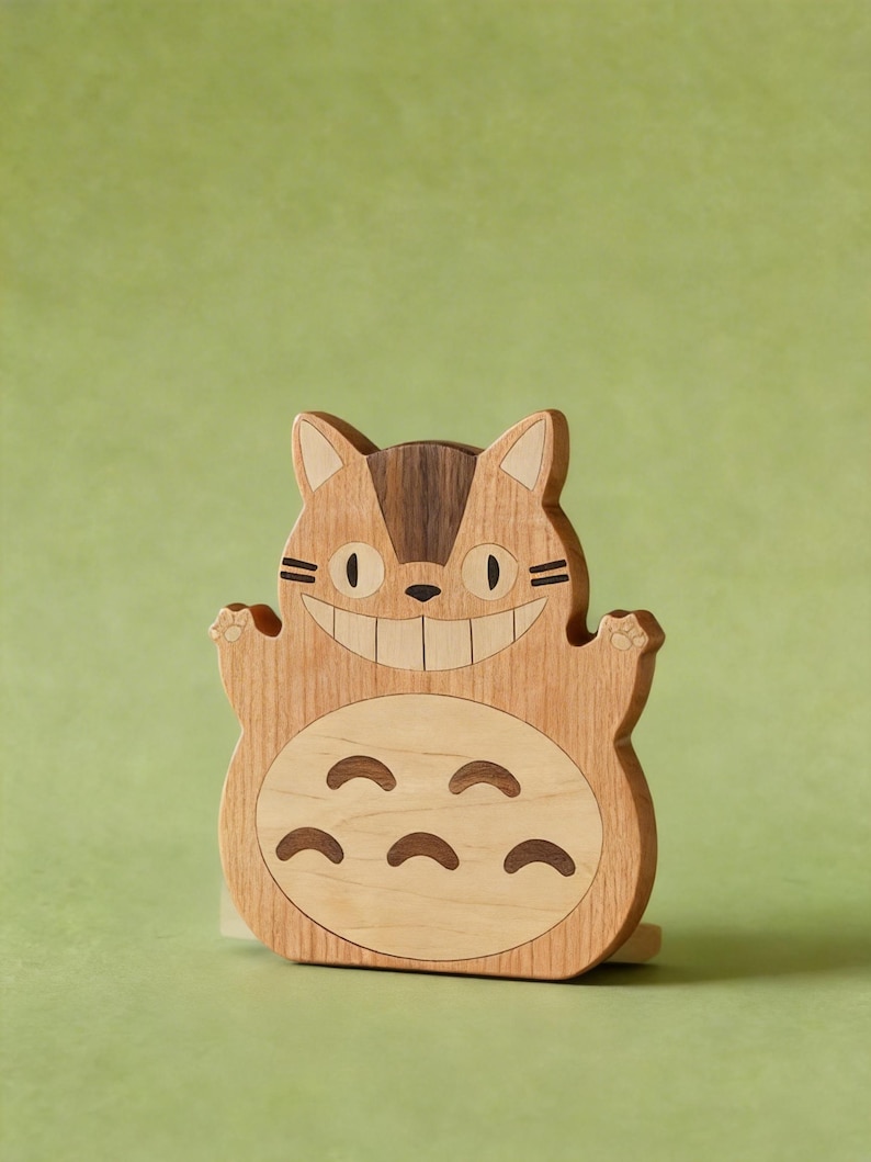 May include: Wooden Totoro figure with a smiling face, paws, and belly markings. The figure is made of light and dark wood tones, with a light green background.