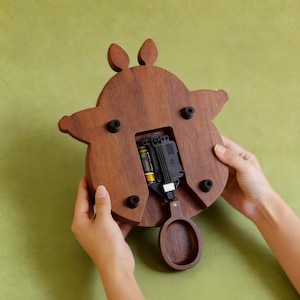 May include: A wooden clock with a brown finish, shaped like a stylized animal. The clock's back is visible, revealing the battery compartment and pendulum. The clock is held by two hands against a green background.