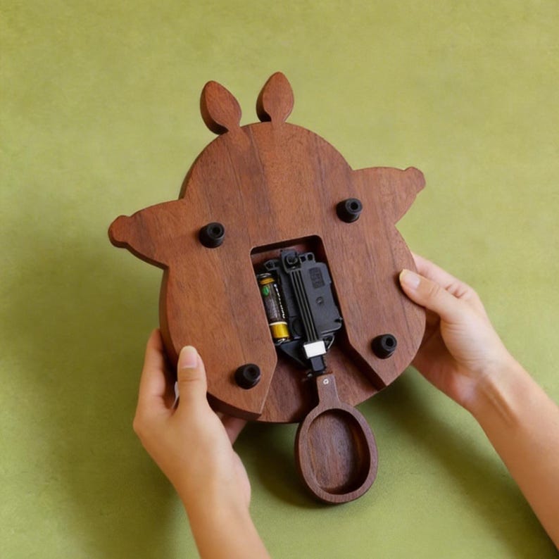May include: A wooden clock with a brown finish, shaped like a stylized animal. The clock's back is visible, revealing the battery compartment and pendulum. The clock is held by two hands against a green background.