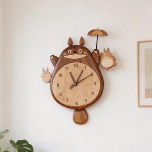 May include: A wooden wall clock shaped like a large brown creature with pointed ears, holding a small umbrella. The clock face is light wood with black numerals from 1 to 12 and dark hands. Two smaller creatures are attached to the sides.