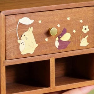 K&ouml;nnte beinhalten: Eine Holz-Aufbewahrungsbox mit einer Schublade mit skurrilen Tier- und Sterneinlagen. Die Schublade hat einen goldfarbenen Knopf. Die Box ist aus warm get&ouml;ntem Holz gefertigt und hat zwei offene F&auml;cher darunter.