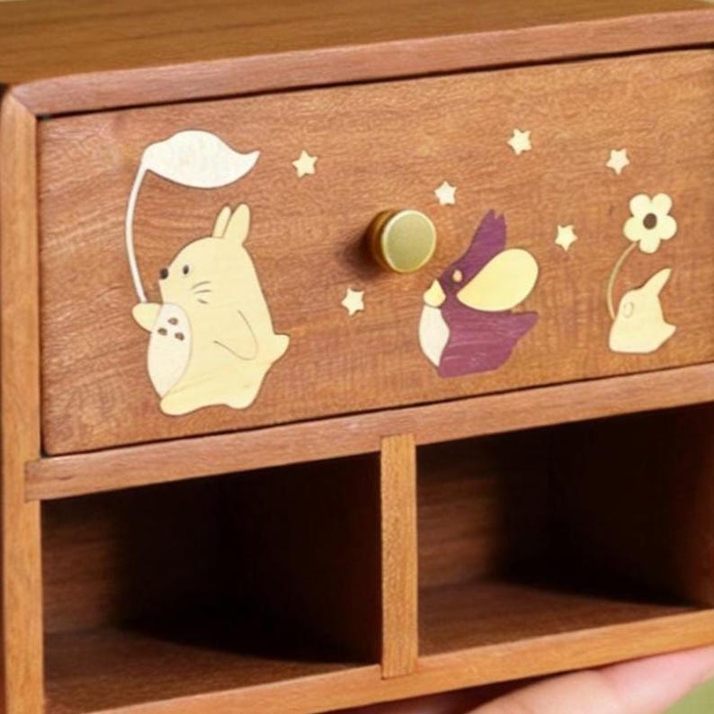 K&ouml;nnte beinhalten: Eine Holz-Aufbewahrungsbox mit einer Schublade mit skurrilen Tier- und Sterneinlagen. Die Schublade hat einen goldfarbenen Knopf. Die Box ist aus warm get&ouml;ntem Holz gefertigt und hat zwei offene F&auml;cher darunter.