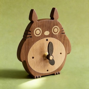 May include: A wooden clock in the shape of a cartoon character, featuring a light brown face and a darker brown body. The clock has gold-colored accents and black clock hands. The clock is approximately 6 inches tall.
