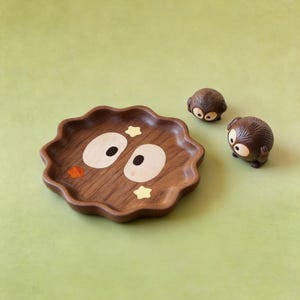 May include: A wooden decorative tray with a wavy edge, featuring a cartoon face design with large oval eyes and star accents. Two small, round, brown figurines with similar eye designs are next to the tray. The background is a solid light green.
