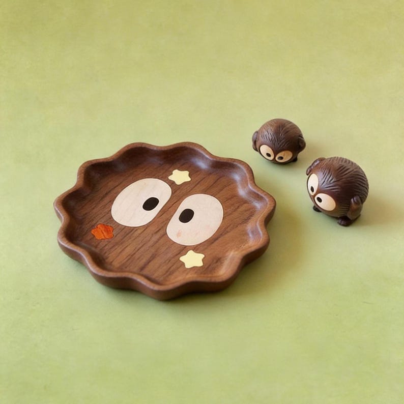 May include: A wooden decorative tray with a wavy edge, featuring a cartoon face design with large oval eyes and star accents. Two small, round, brown figurines with similar eye designs are next to the tray. The background is a solid light green.