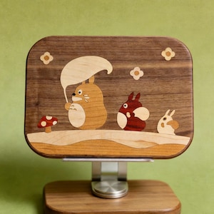 May include: A rectangular wooden stand featuring an inlaid marquetry design of three cartoon creatures. The largest creature holds a light-colored leaf umbrella, accompanied by a smaller red creature and a tiny white one, with a mushroom nearby.