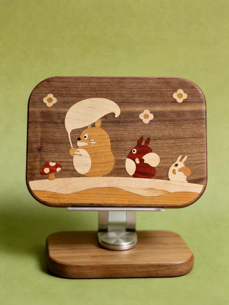 May include: A rectangular wooden stand featuring an inlaid marquetry design of three cartoon creatures. The largest creature holds a light-colored leaf umbrella, accompanied by a smaller red creature and a tiny white one, with a mushroom nearby.