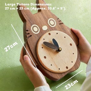 May include: Wooden Totoro-shaped clock, wall-ready. The clock features a light wood clock face with dark hands and a dark wood Totoro outline. Dimensions are approximately 10.6" x 9". The clock can be wall-mounted or displayed on a tabletop.