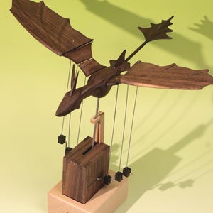 May include: A mechanical wooden dragon automaton featuring dark walnut wings and body, suspended by thin metal wires above a rectangular dark wood housing. The entire mechanism is mounted on a light-colored, smooth wooden base with a small brass knob.