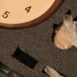 May include: A light-coloured wooden clock face with dark brown numerals 4, 5, and 6, partially encased in dark wood. The clock is nestled in dark grey protective foam packaging with cutouts containing small components wrapped in clear plastic.