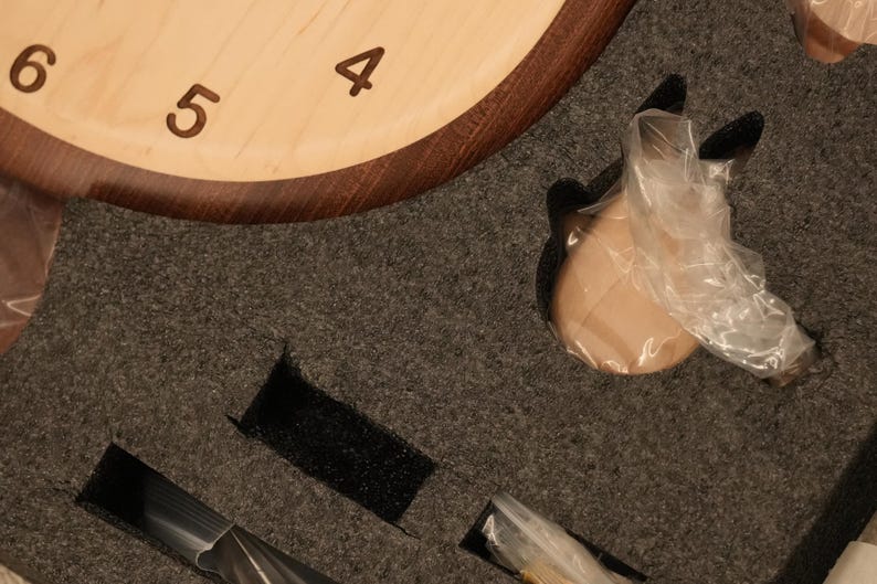 May include: A light-coloured wooden clock face with dark brown numerals 4, 5, and 6, partially encased in dark wood. The clock is nestled in dark grey protective foam packaging with cutouts containing small components wrapped in clear plastic.