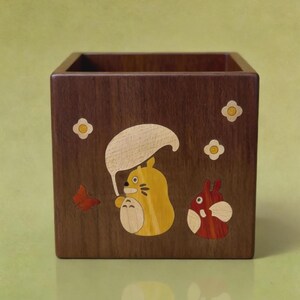 May include: A wooden square box with a dark brown finish. The box is decorated with inlaid wood designs of a yellow character with a white umbrella, a red character, a red butterfly, and white flowers.