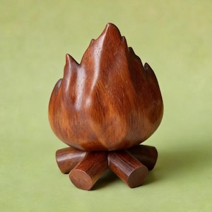 May include: A handcrafted wooden campfire sculpture. The sculpture is made of dark brown wood and features a flame-like shape atop a base of wooden logs. The wood grain is visible, adding to the natural aesthetic. The campfire is set against a green background.