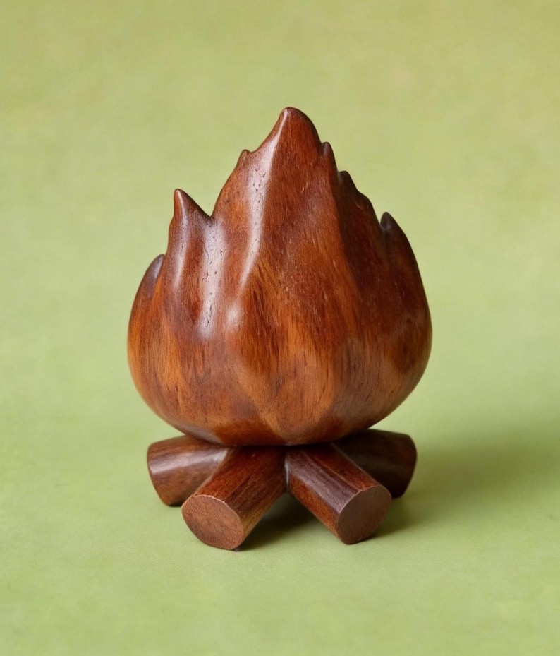 May include: A handcrafted wooden campfire sculpture. The sculpture is made of dark brown wood and features a flame-like shape atop a base of wooden logs. The wood grain is visible, adding to the natural aesthetic. The campfire is set against a green background.