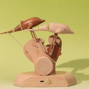 Peut inclure: Une sculpture mécanique en bois représentant deux dauphins, l'un brun foncé et l'autre beige clair, fixés sur un socle en bois clair. La structure cinétique comprend des bras articulés, des engrenages et un petit bouton métallique circulaire.