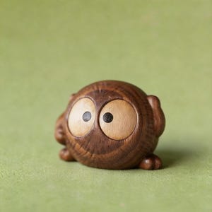 May include: A small, wooden owl figurine with large, light-colored eyes and dark pupils. The owl is carved from a dark brown wood and has a rounded body with small feet. The figurine is set against a green background.