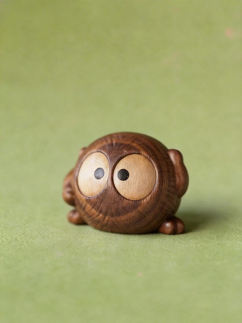 May include: A small, wooden owl figurine with large, light-colored eyes and dark pupils. The owl is carved from a dark brown wood and has a rounded body with small feet. The figurine is set against a green background.