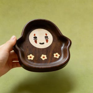May include: A dark brown wooden tray with a No-Face design in the center and three small flower designs. The No-Face design has a white circle with black eyes and a red mouth. The tray is held by a hand against a green background.