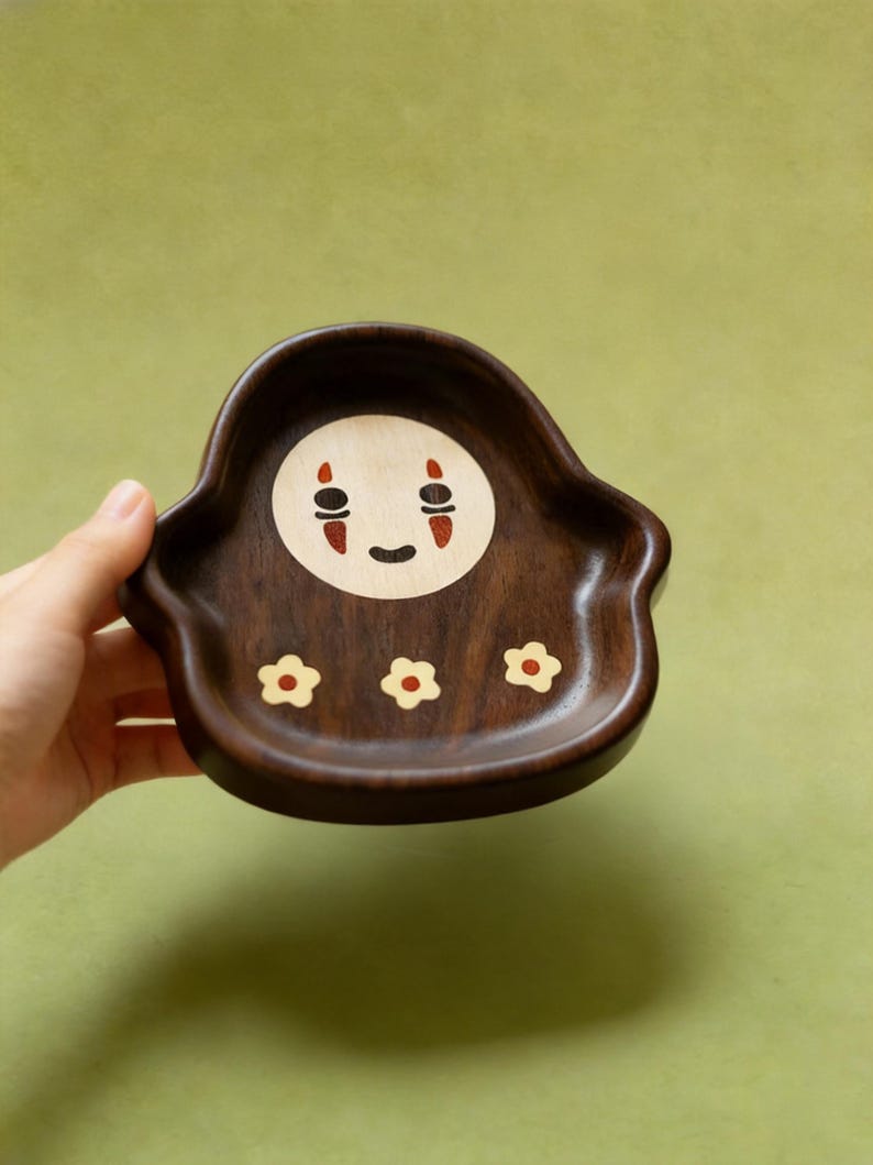 May include: A dark brown wooden tray with a No-Face design in the center and three small flower designs. The No-Face design has a white circle with black eyes and a red mouth. The tray is held by a hand against a green background.