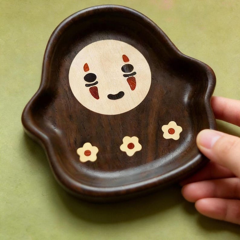 May include: A handcrafted wooden tray with a dark brown finish. The tray features a central circular design with a smiling face and three small flower accents. The face has black eyes, a black mouth, and red accents.