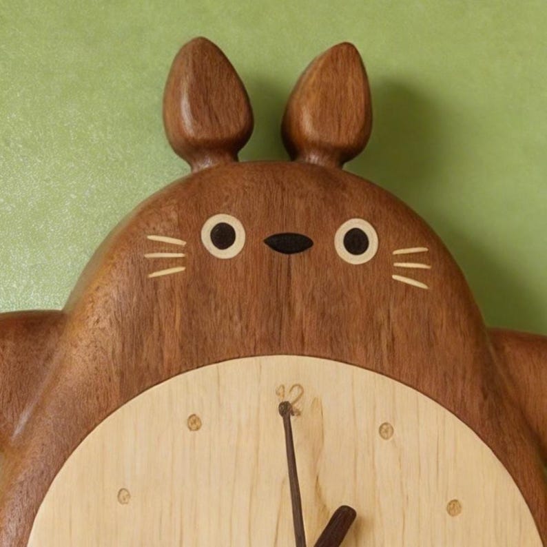 May include: Wooden wall clock featuring a cartoon character design with a light wood face and a dark brown wooden body. The clock has two round eyes, a small black nose, and three whiskers on each side. The clock hands are black.