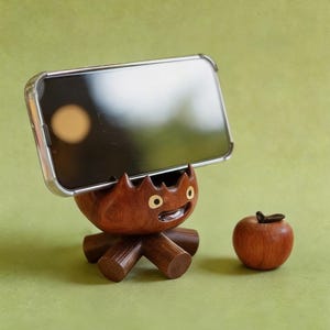 May include: A wooden phone stand shaped like a cartoon character with a smiling face and a small wooden apple. The stand is holding a phone with a clear case. The stand and apple are brown, set against a green background.